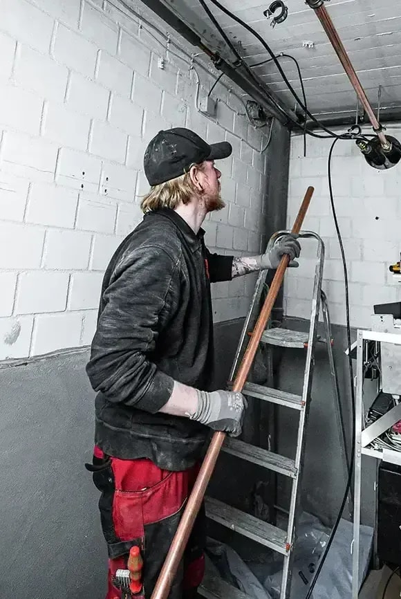 Heizungsmonteur verlegt Kupferrohr an einer Leiter im Kellerraum bei der Heizungsinstallation